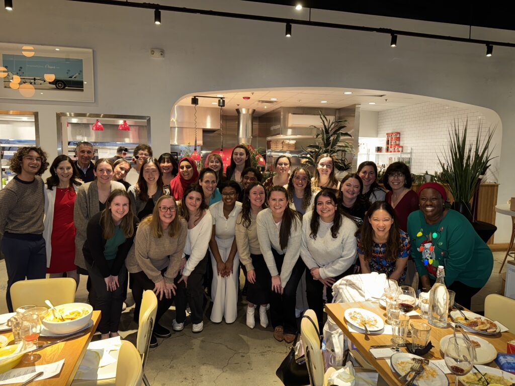 More than two dozen people pose for camera in large kitchen after a group meal.