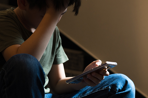 Young preteen/teenage boy sit on stair at home, cry and cover his face with hand, other hand hold smartphone, feeling alone, stressed, frustrated, overwhelmed, depressed. Cyber bullying concept.